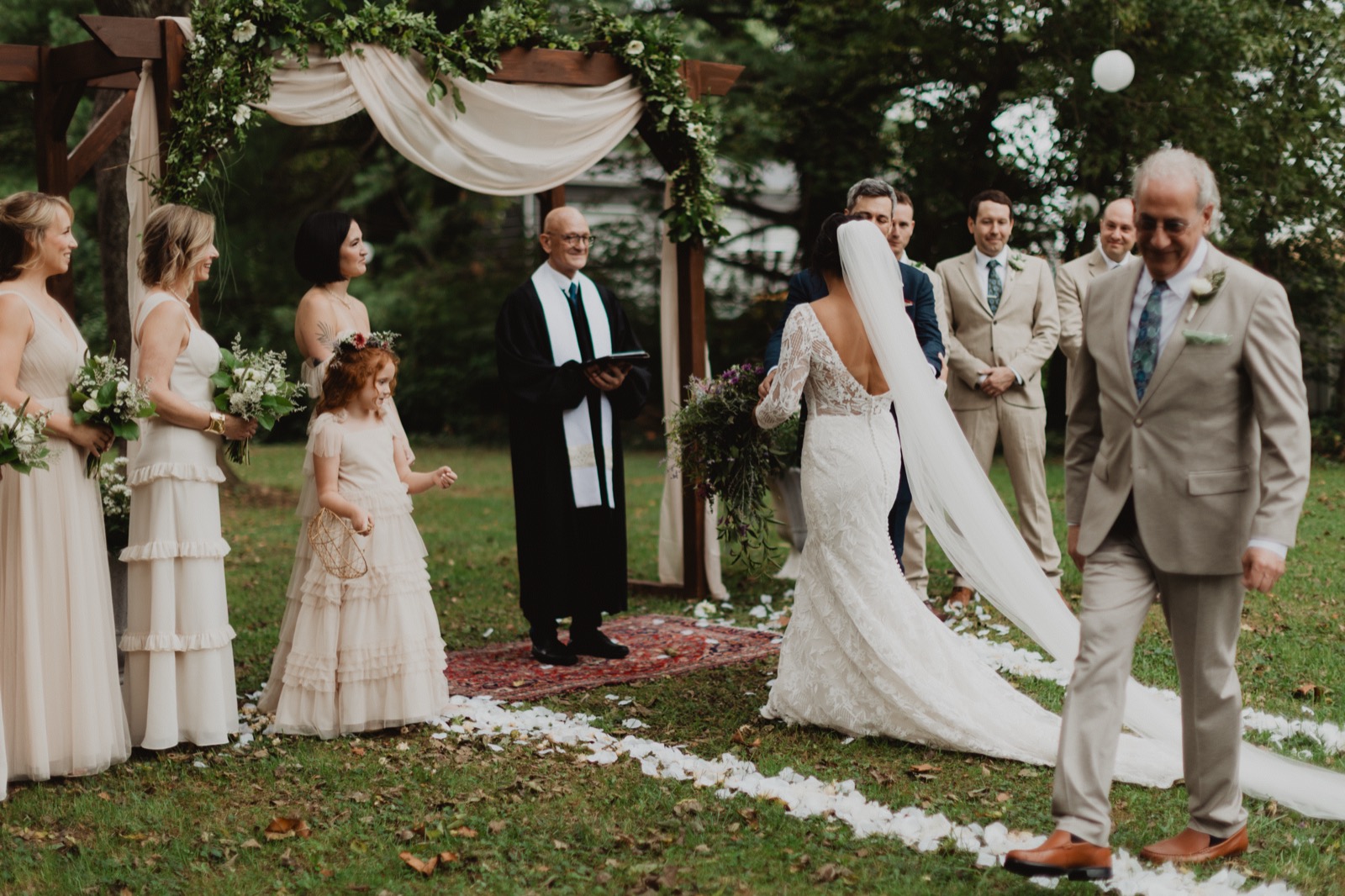 A bride walking the last of a petal-strewn aisle on a lawn toward a draped timber arbour, bridesmaids and a flower girl watching from one side and the officiant waiting beneath the greenery
