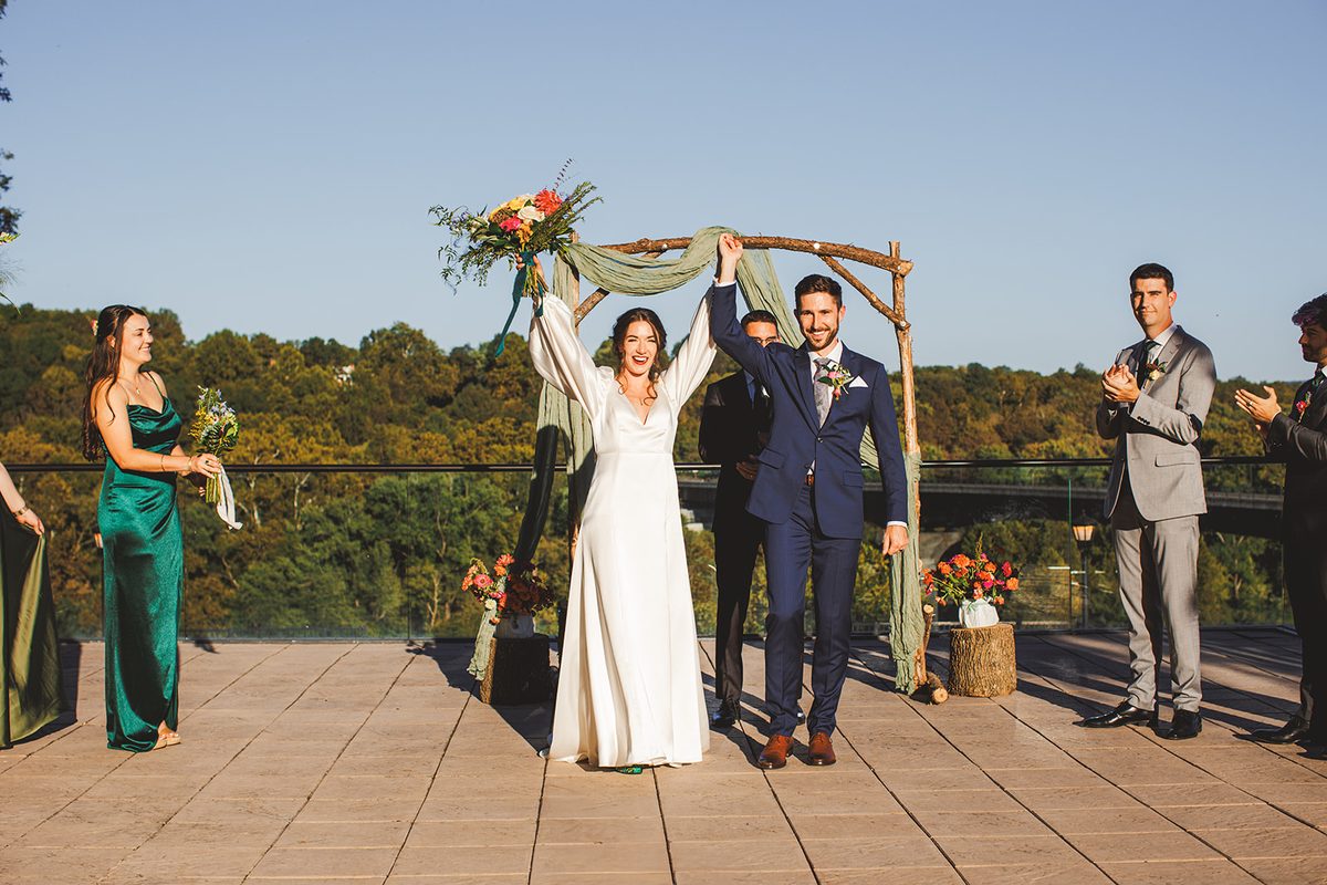 Andrea raising her bouquet and Patrick raising their joined hands beneath the arch just after being pronounced married