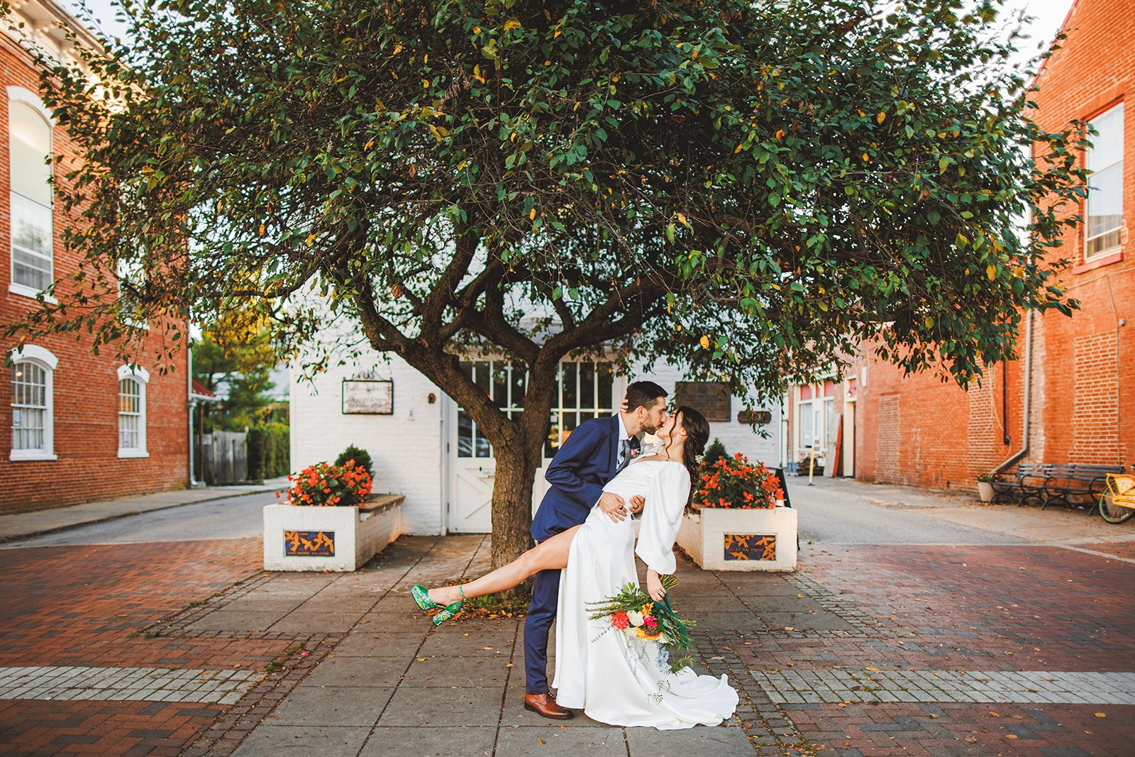 A couple in a dramatic dip kiss beneath a large canopy tree on a brick town street, one green heel kicked up