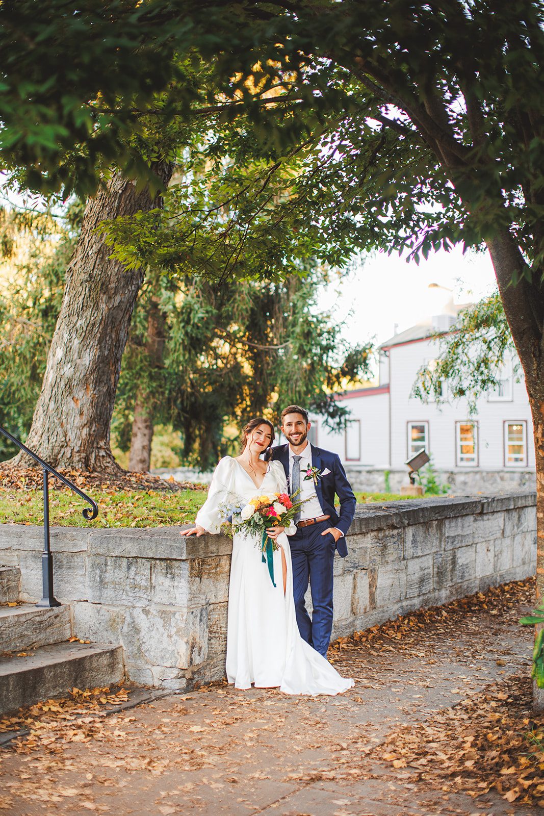 Andrea and Patrick leaning together against the stone wall beneath tall trees, fallen leaves across the path and the inn glowing white behind them