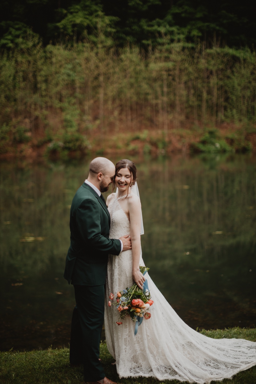 A couple standing at the edge of a still pond in the woods, foreheads close and both laughing, her lace train spread across the grass and the treeline mirrored in the water behind them