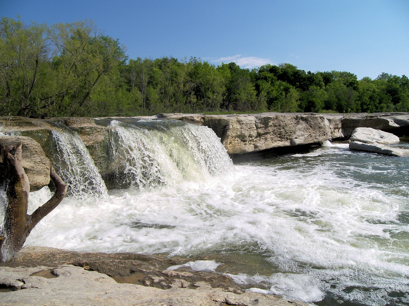 Onion Creek pouring in a wide sheet over the limestone ledge at McKinney Falls, white water breaking into the pool below with green woods behind