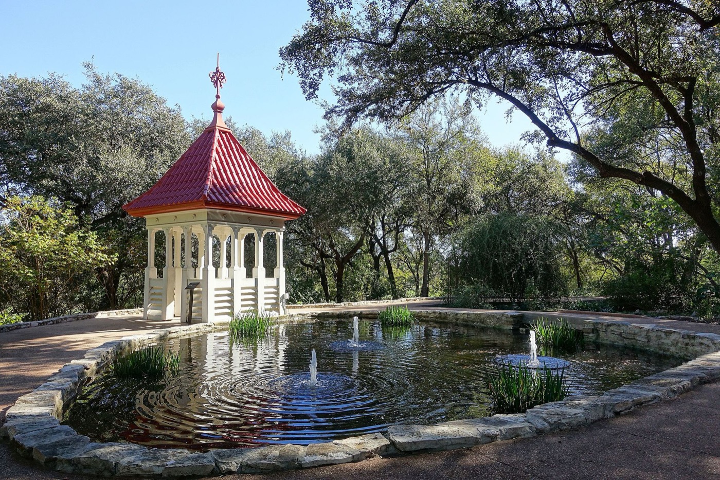 A small red-roofed pagoda on a stone terrace beside a still pond in a botanical garden, live oaks arching overhead and fountains breaking the water