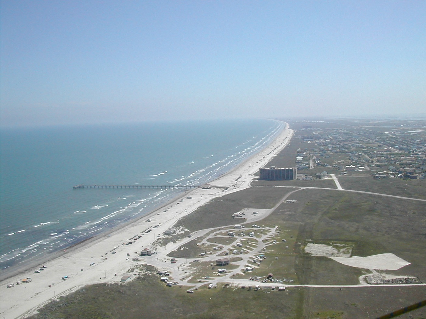 An aerial view along Mustang Island, the long straight beach running away into haze with the Gulf on one side and low coastal town and dunes on the other