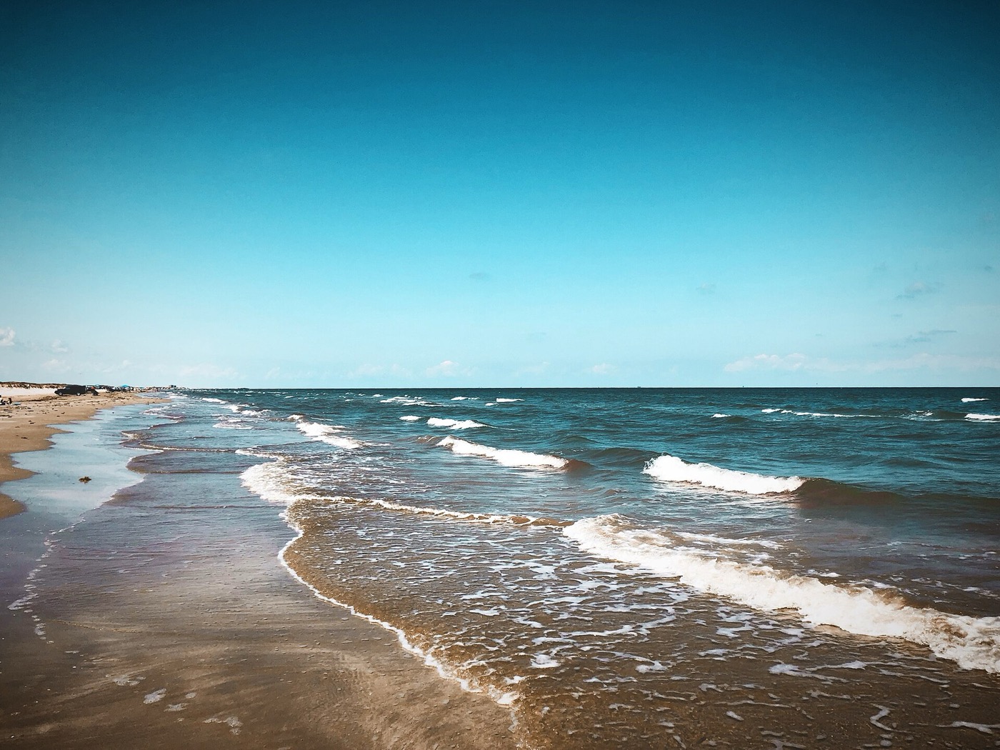 Clear turquoise Gulf water running up a wide flat beach under a pale blue sky, small waves breaking along an empty shoreline