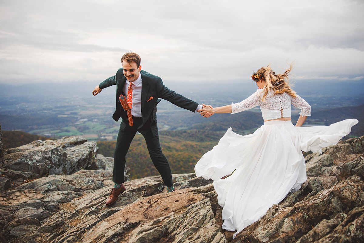 Ryan leading Sara by the hand across the summit boulders, her white skirt blowing sideways in the wind under an overcast sky