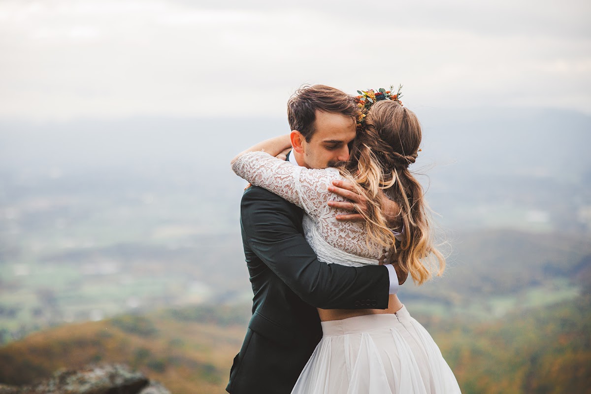 Ryan wrapping Sara in a hug on the summit, her hair and flower crown against his shoulder, the hazy valley floor far below