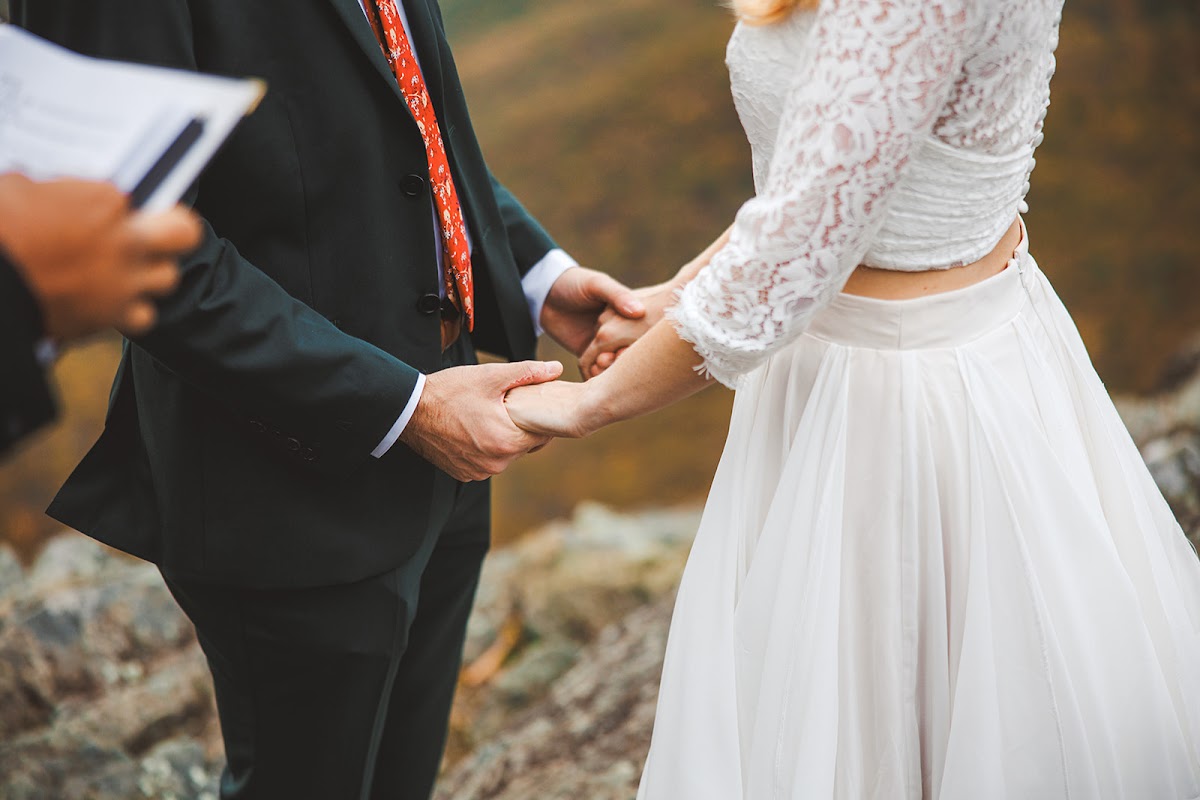Close frame of Sara and Ryan holding both hands during vows, her lace sleeves and flowing skirt against his dark green suit and red floral tie, the officiant's book at the edge of the frame