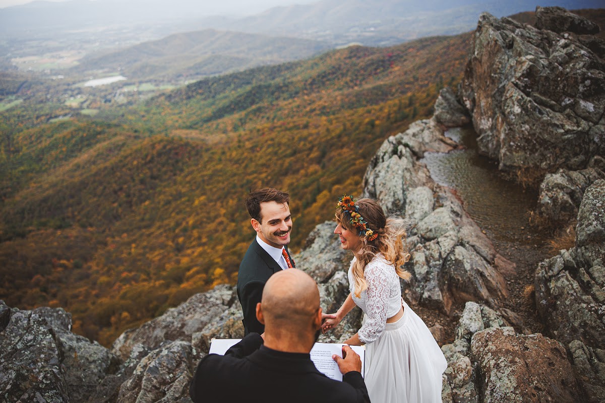 Sara and Ryan holding hands during vows on a rocky overlook in Shenandoah National Park, the officiant reading beside them and the autumn mountainside falling away behind