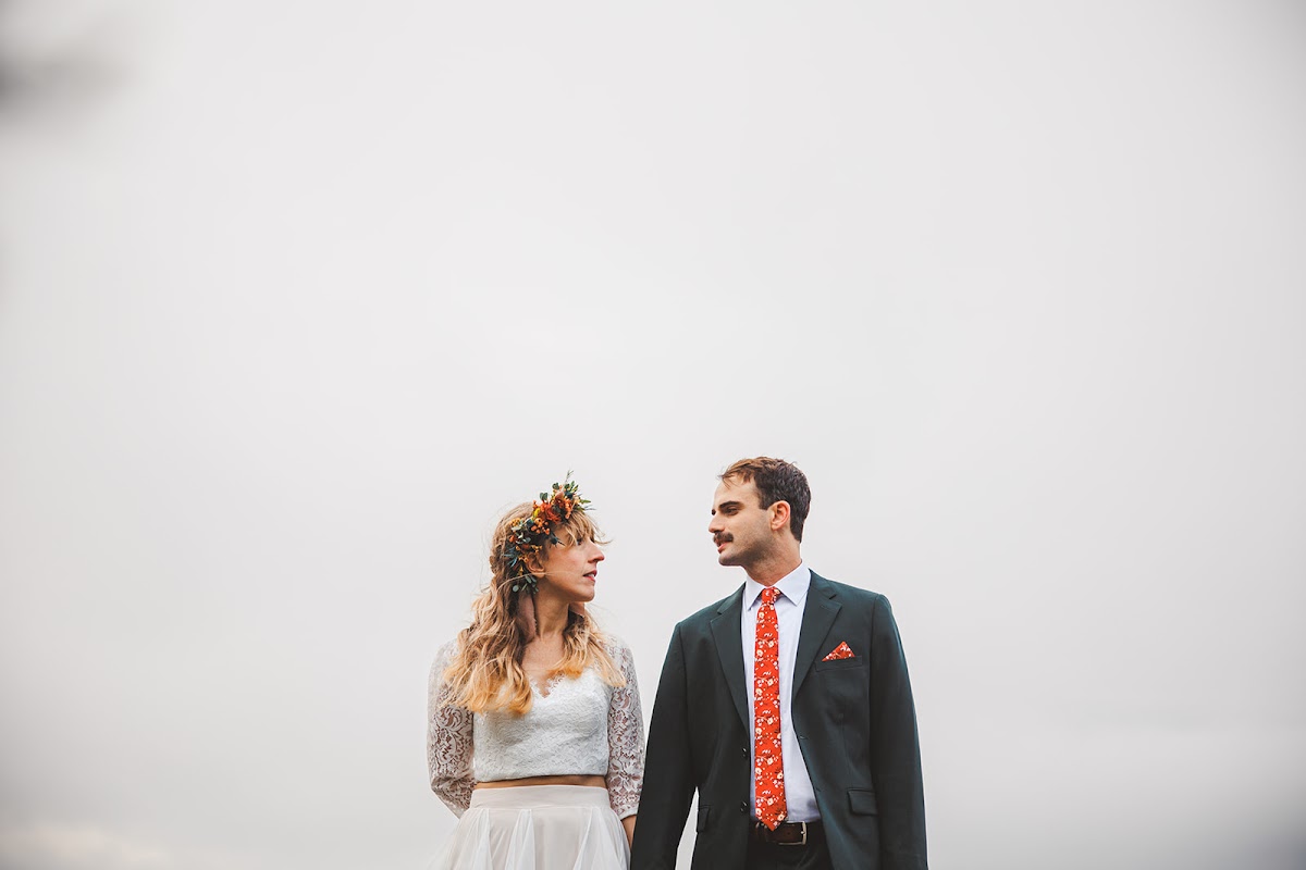 Minimal portrait of Sara in her flower crown and Ryan in a dark green suit with an orange floral tie, holding hands against a white sky