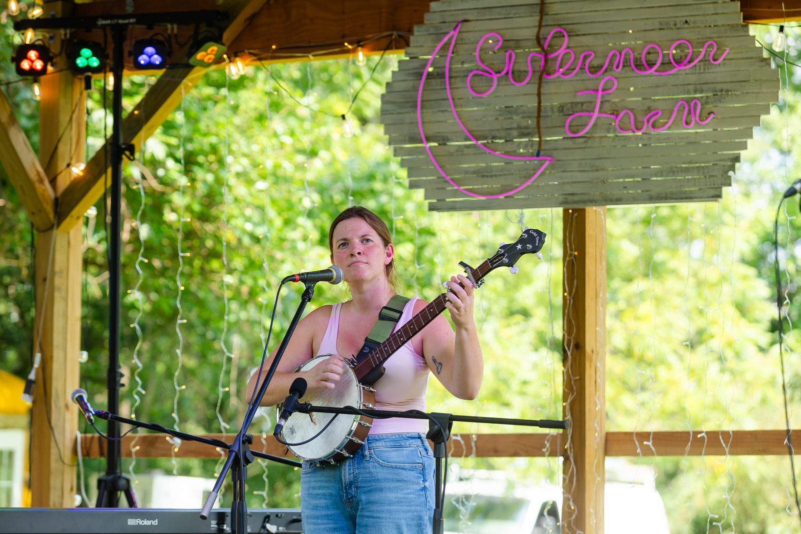 A banjo player singing at the microphone under the pink neon Sistermoon Farm sign on the timber stage at Tomato Jam