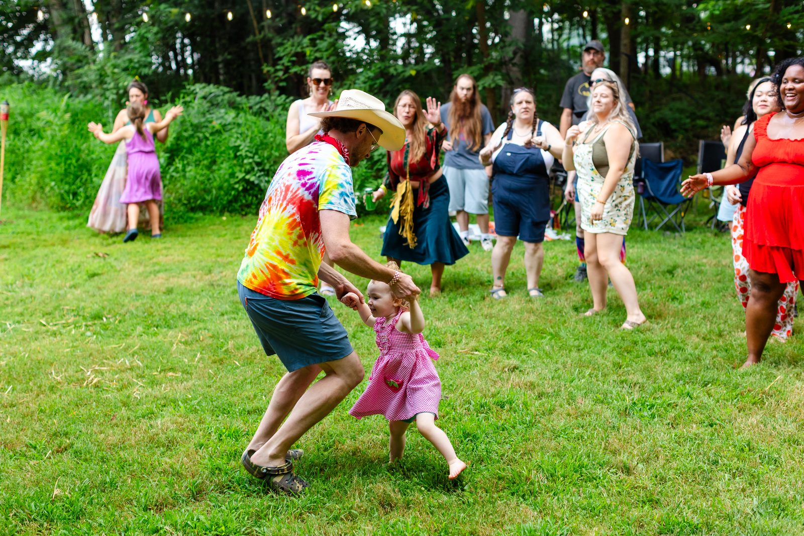 A man in tie-dye and a cowboy hat swinging a little girl in a pink dress by the hands mid-dance