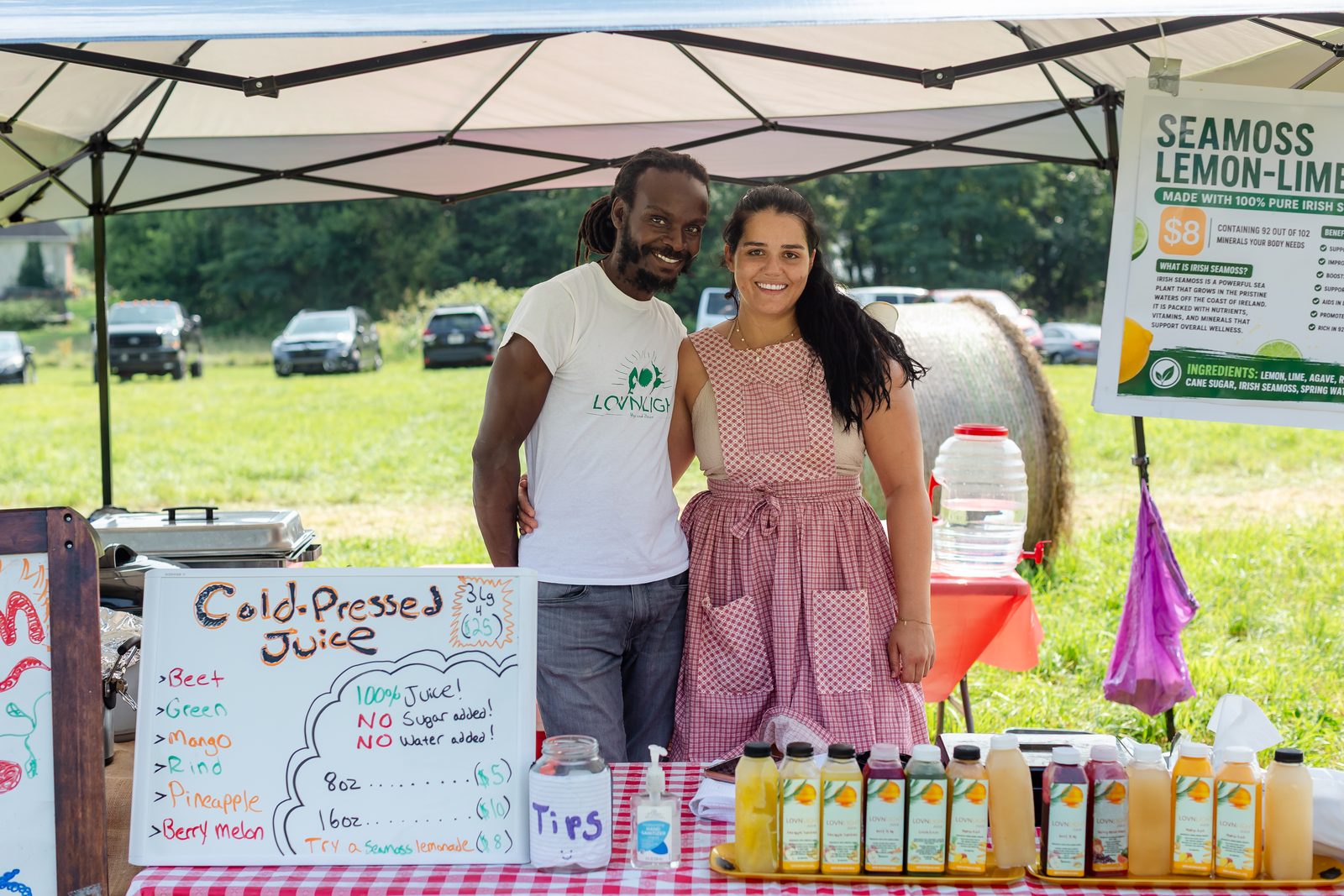 Two vendors standing arm in arm behind their cold-pressed juice stand, bottles lined up on a checkered tablecloth