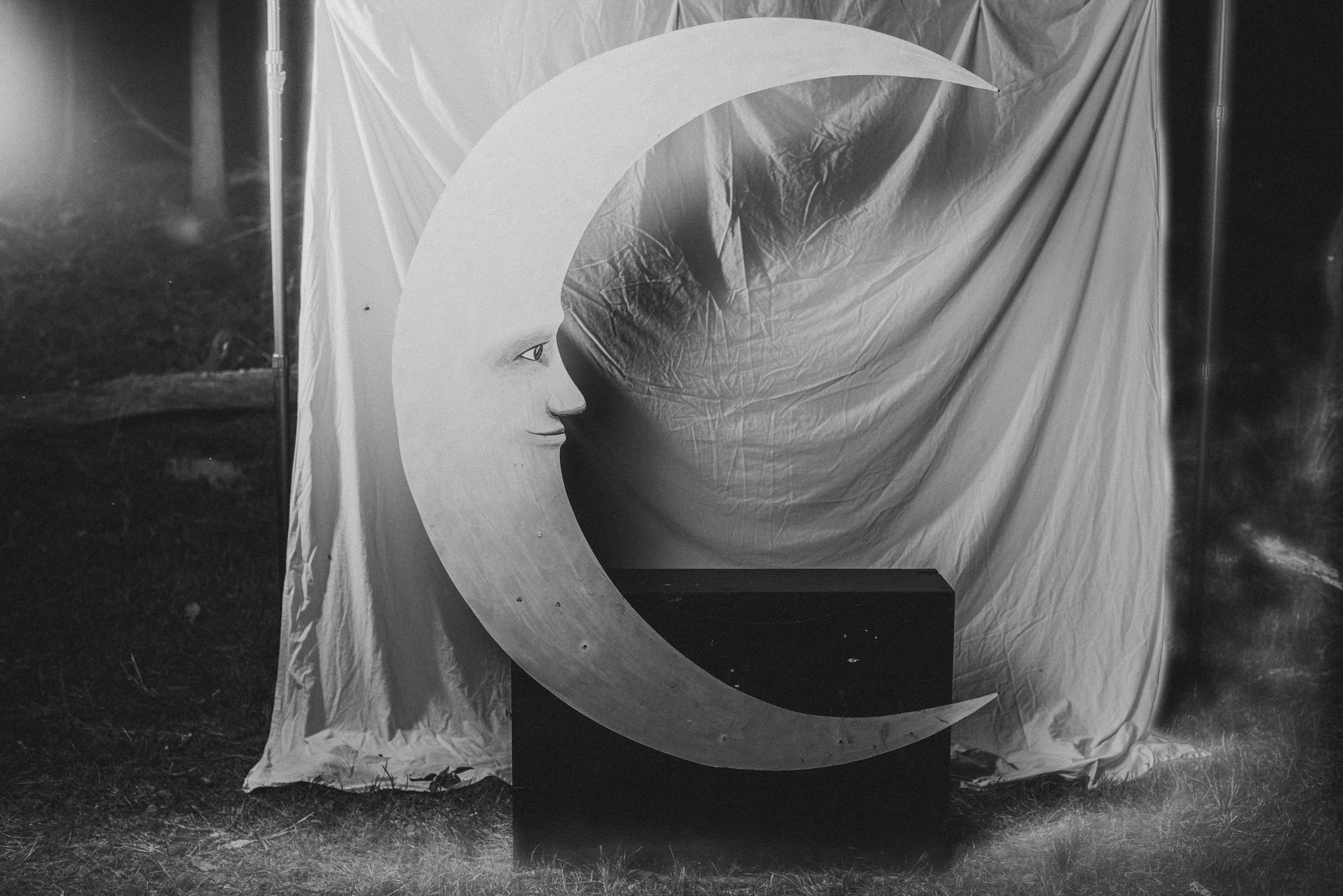The Moon Booth at night at Sistermoon Farm in black and white: a hand-painted wooden crescent moon with a face, a black bench, and a white sheet backdrop