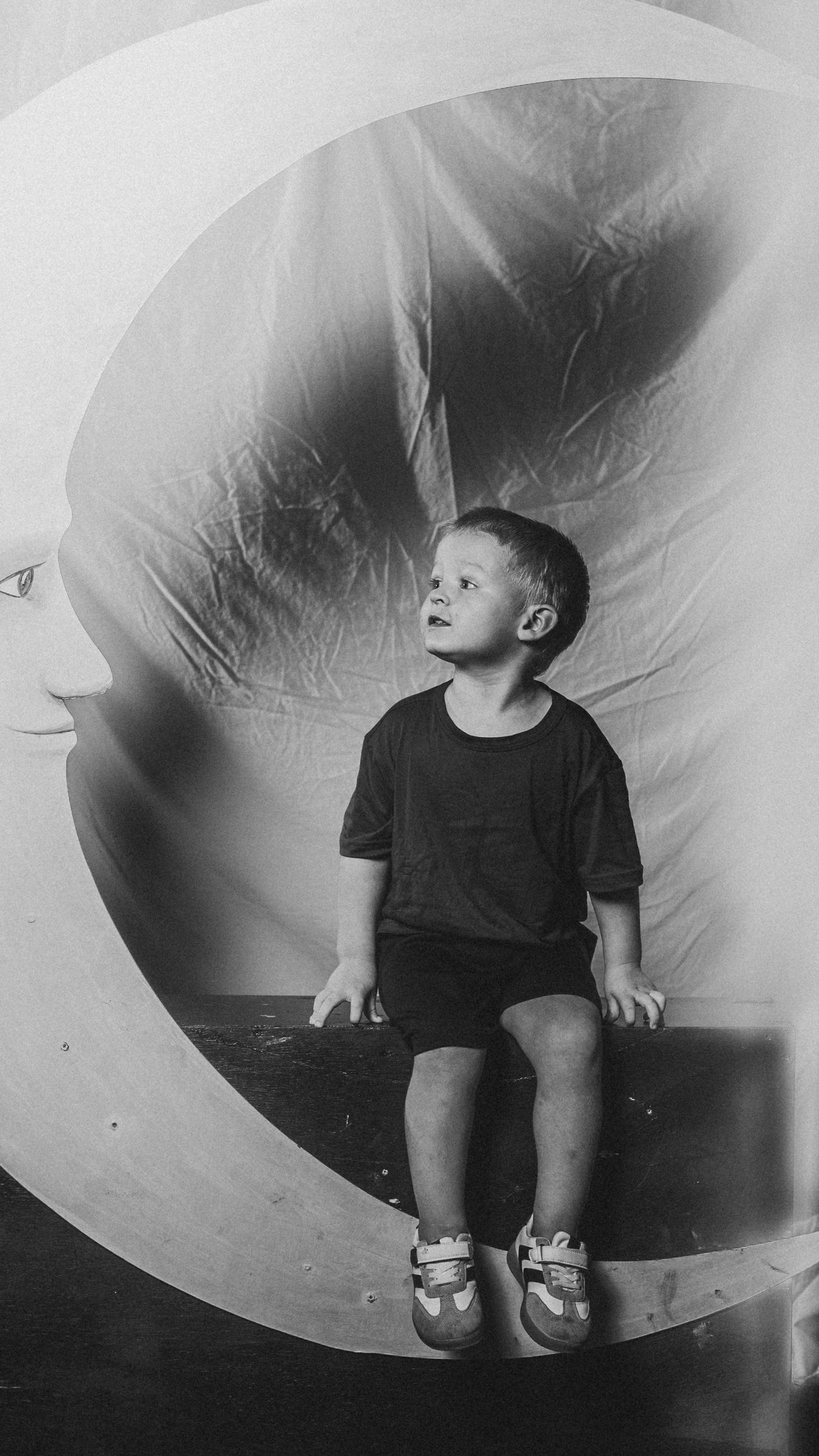 A toddler sitting at the base of the Moon Booth crescent moon, looking up at its painted face, in black and white