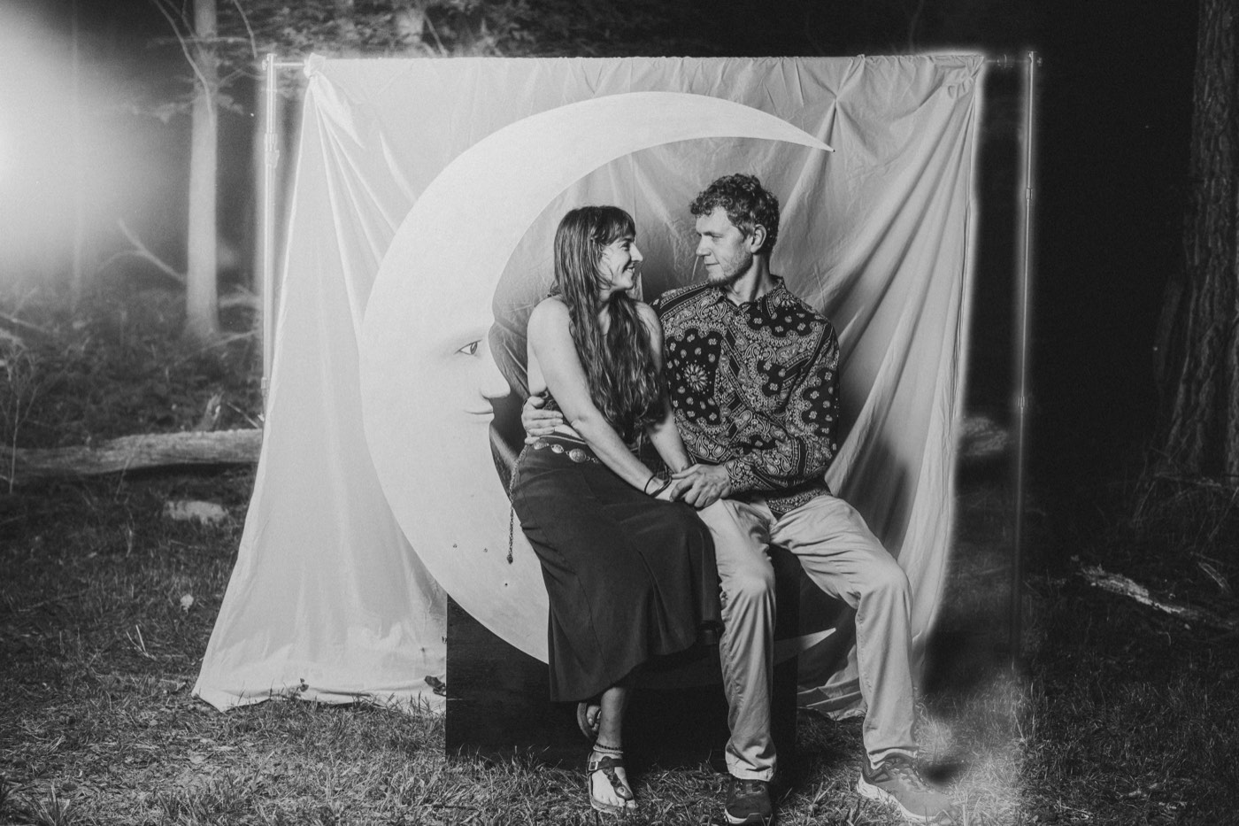 A couple seated together on a plywood crescent moon, photographed in black and white under studio lighting