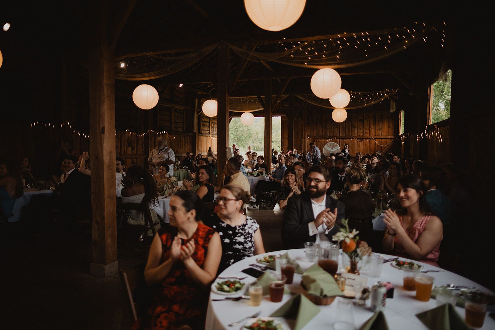 Guests seated at round tables under paper lanterns clap and smile toward the front of the room.