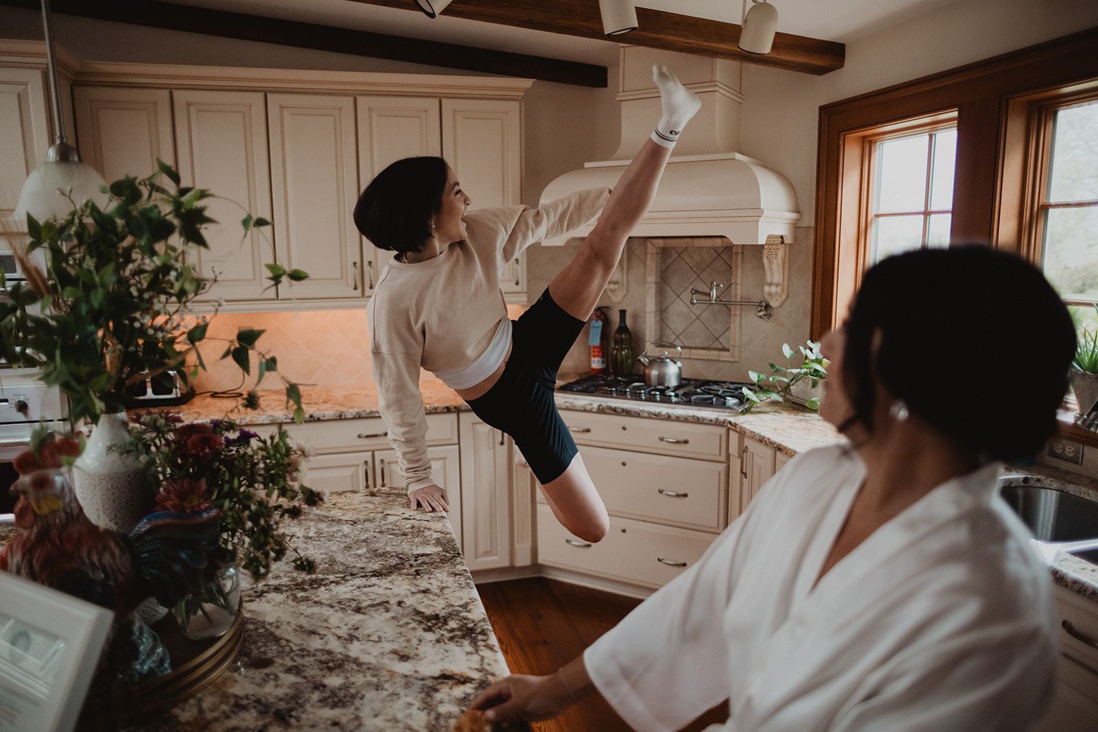 A woman in a sweatshirt and shorts kicks one leg high beside a kitchen island while another woman in a white robe watches from across the counter.