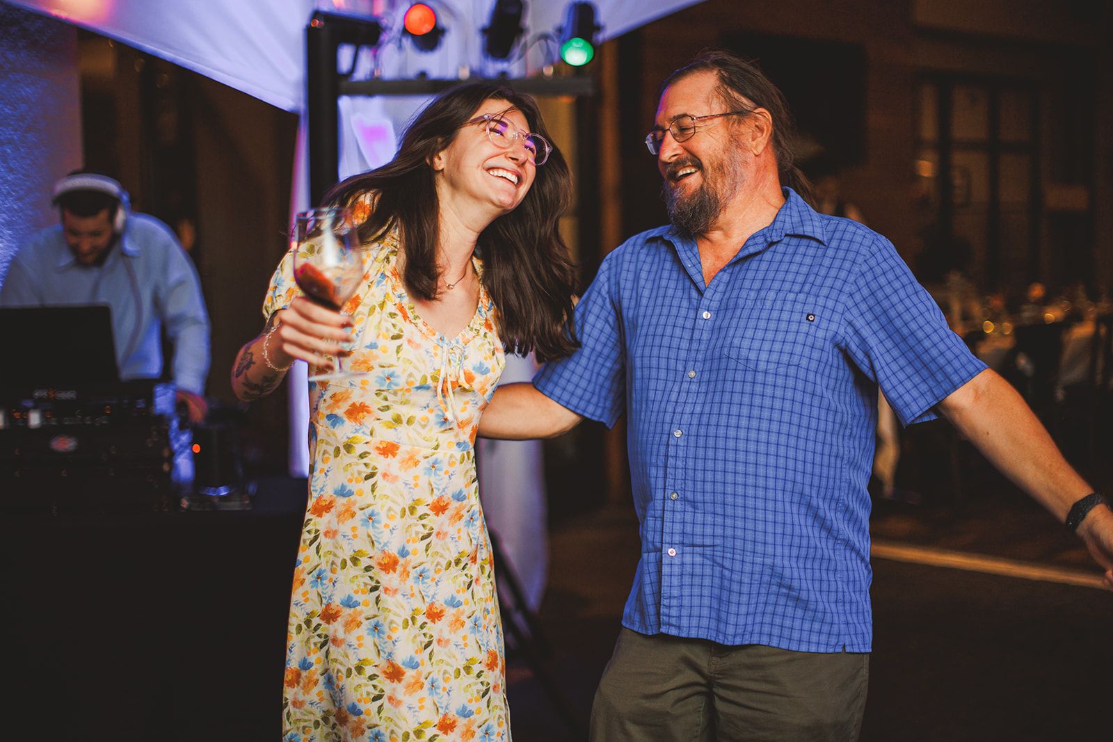 A woman in a floral dress holds a glass of wine and laughs beside an older man in a blue checked shirt as the two dance in front of the DJ booth.