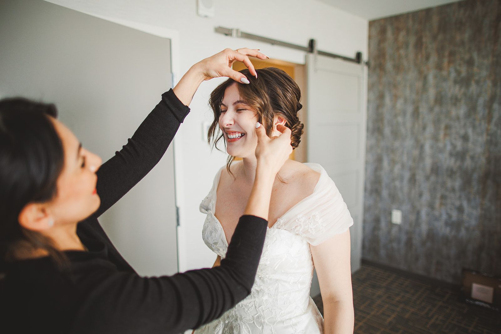 A woman in black uses both hands to settle the bride's hair while the bride laughs with her eyes closed.