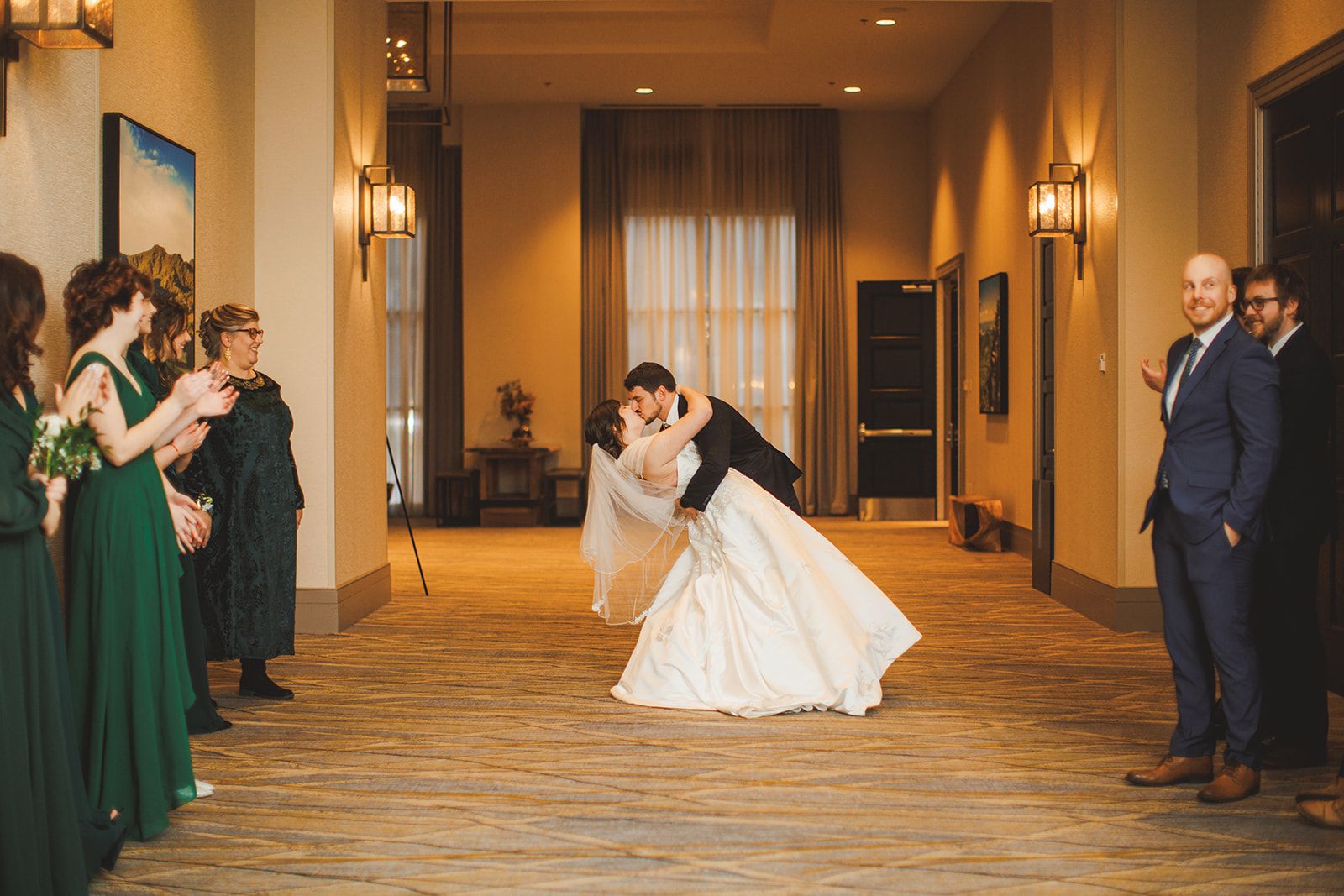 A small group of bridesmaids and guests watches and claps from either side of a warm, empty hallway as the groom dips the bride into a kiss.