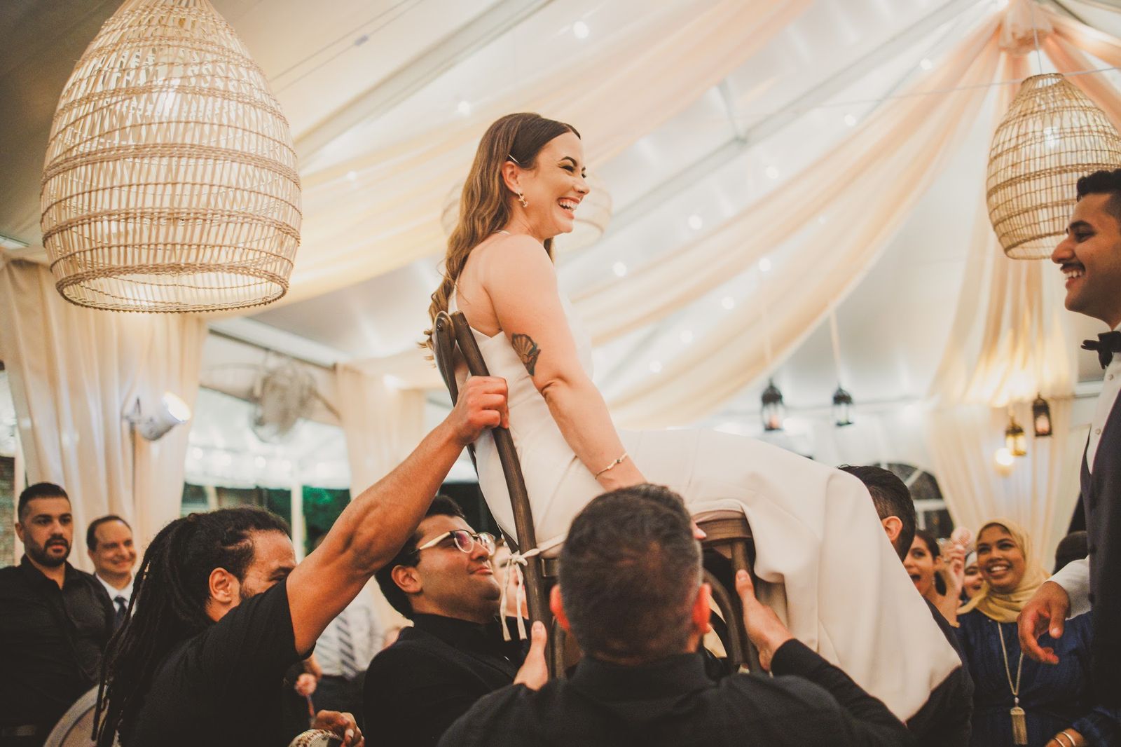 Guests lift the bride overhead on a chair as she laughs and looks down at them.