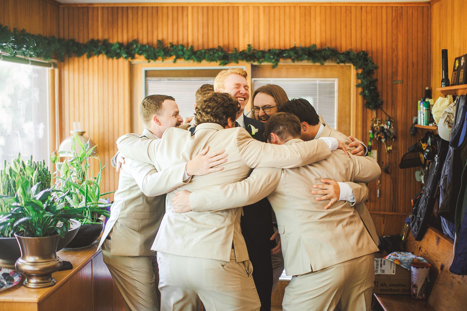 A group of men in tan and dark suits pile into a hug in a wood-paneled room while one of them grins over the top of the huddle.