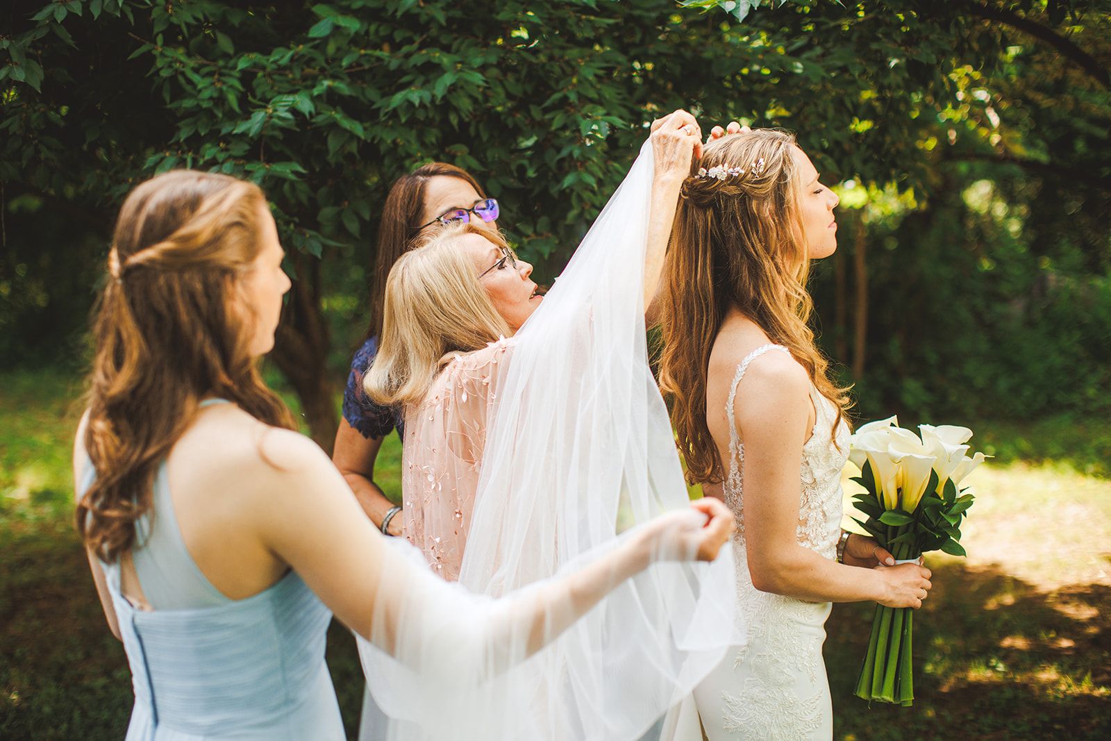 A woman in a pink dress reaches up with both hands to settle a veil into the bride's hair while two other women stand close and the bride holds her calla lily bouquet.
