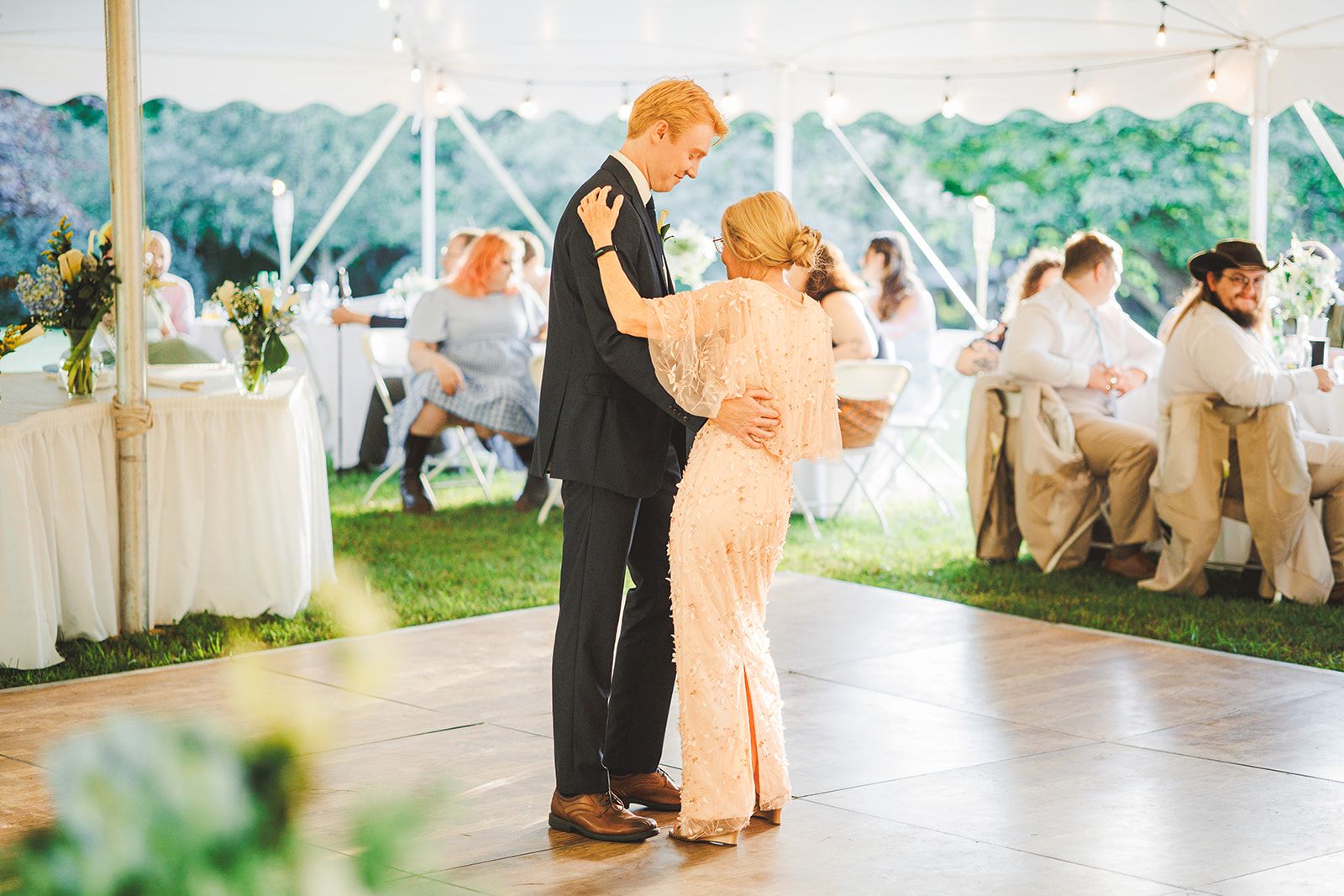 A groom in a black suit dances with an older woman in a blush gown on an empty dance floor under a tent while guests sit at tables behind them.