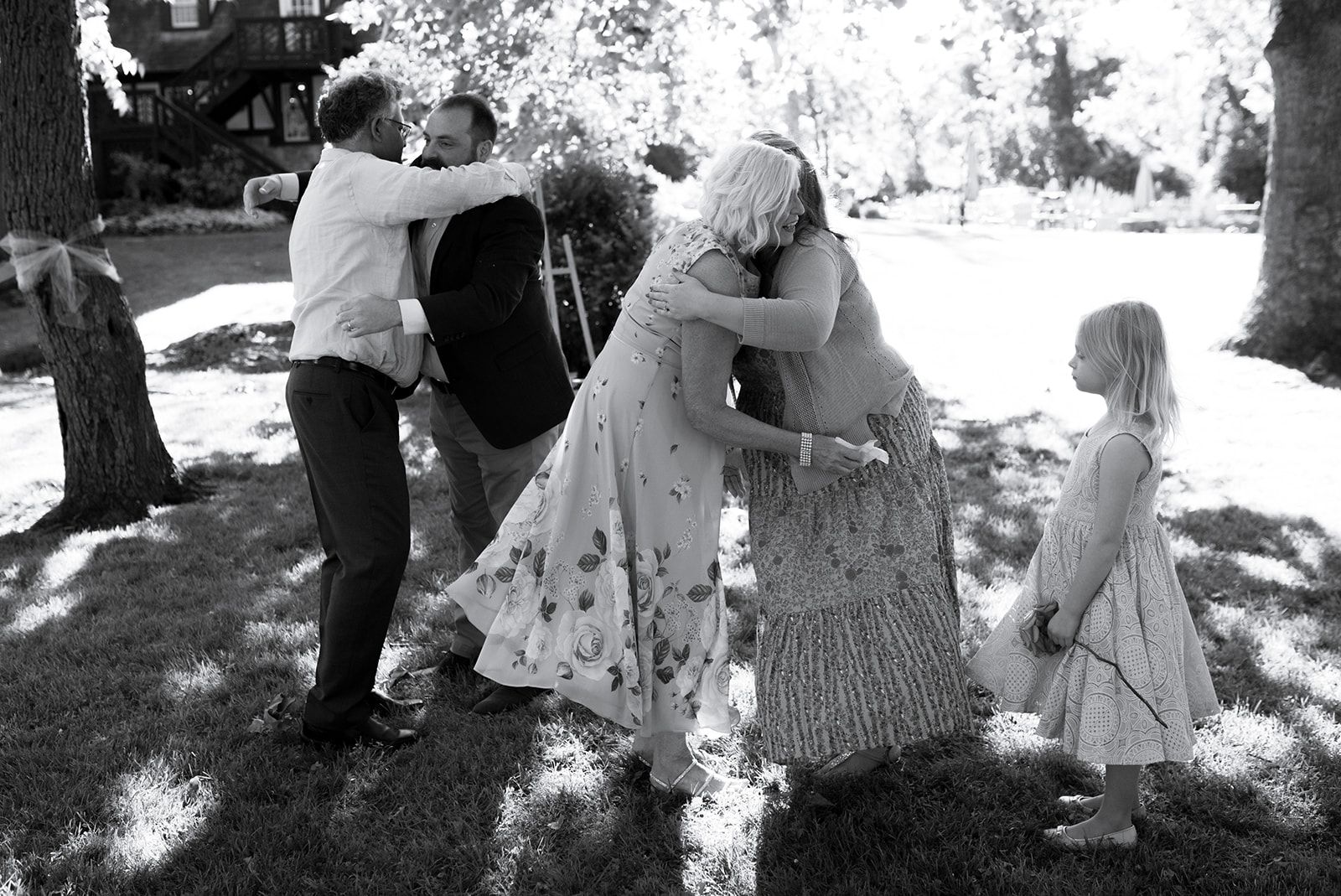 Two pairs of people hug at the same moment on a shaded lawn while a young girl stands watching.