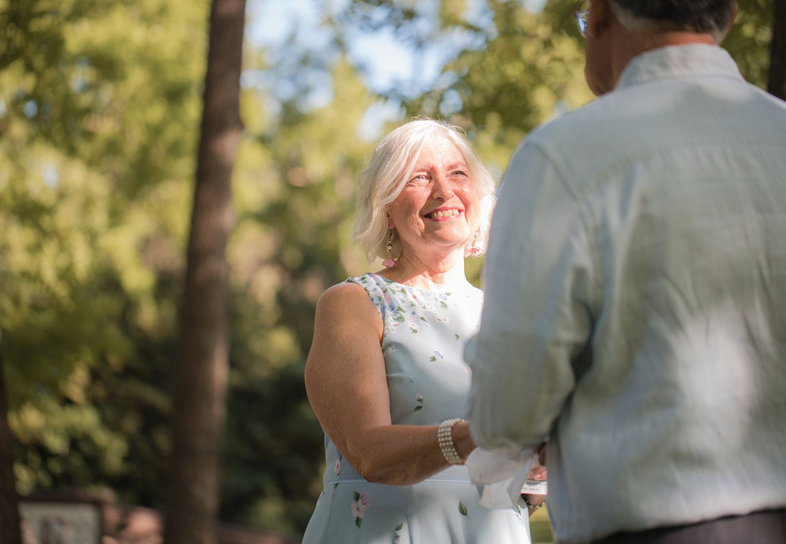 A woman in a pale blue dress beams up at a man whose back is to the camera, sunlit trees behind them.