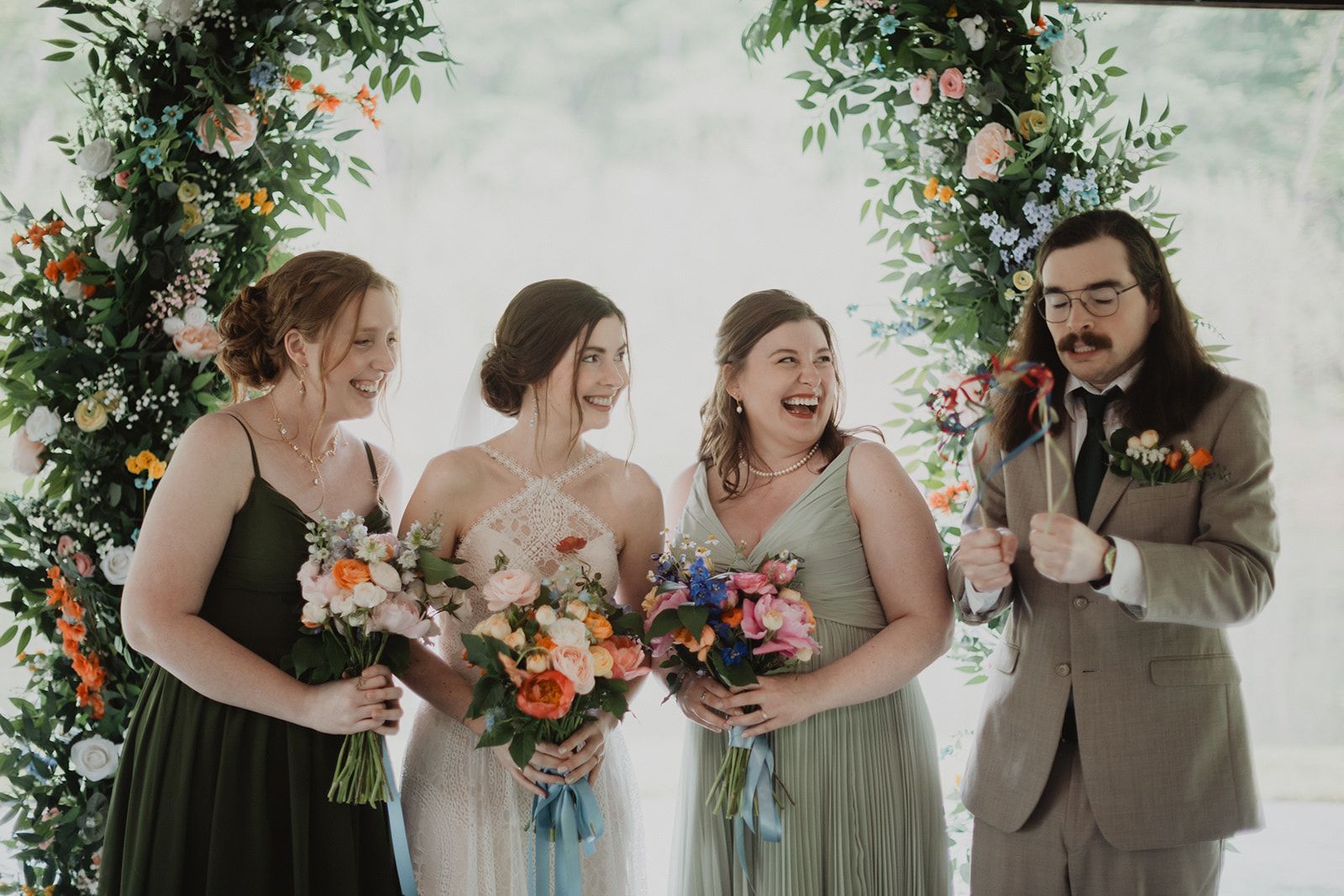 The bride stands between two attendants in green dresses holding bouquets, one of them laughing hard, while a man in a tan suit holding ribbon wands grins at the end of the line.