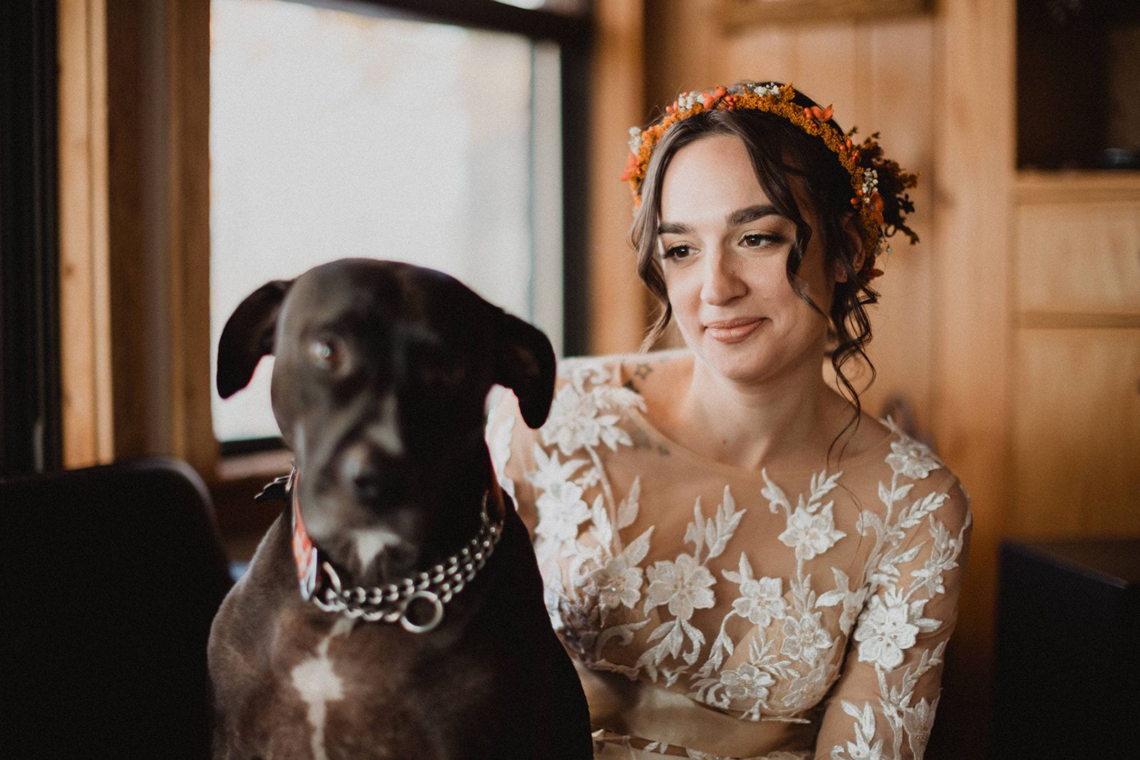 A bride in a lace dress and flower crown sits close beside a large black dog, looking off with a soft smile while the dog faces the camera.