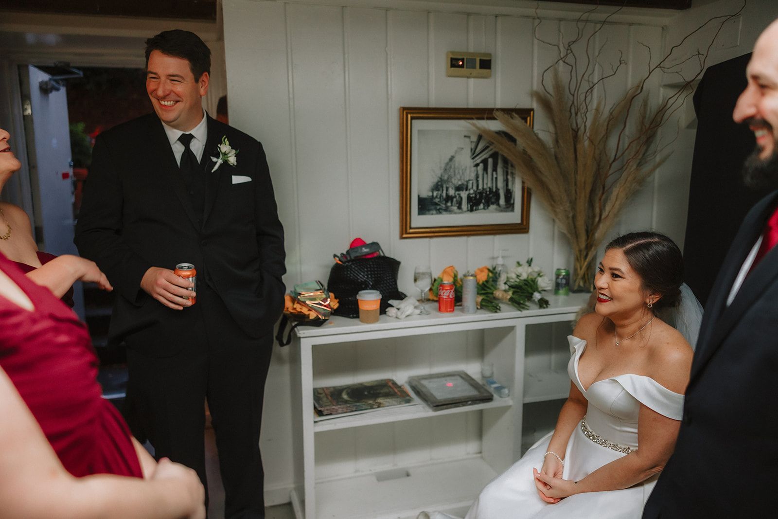 A laughing groom in a black suit stands holding a drink can while the seated bride looks over at him and smiles.