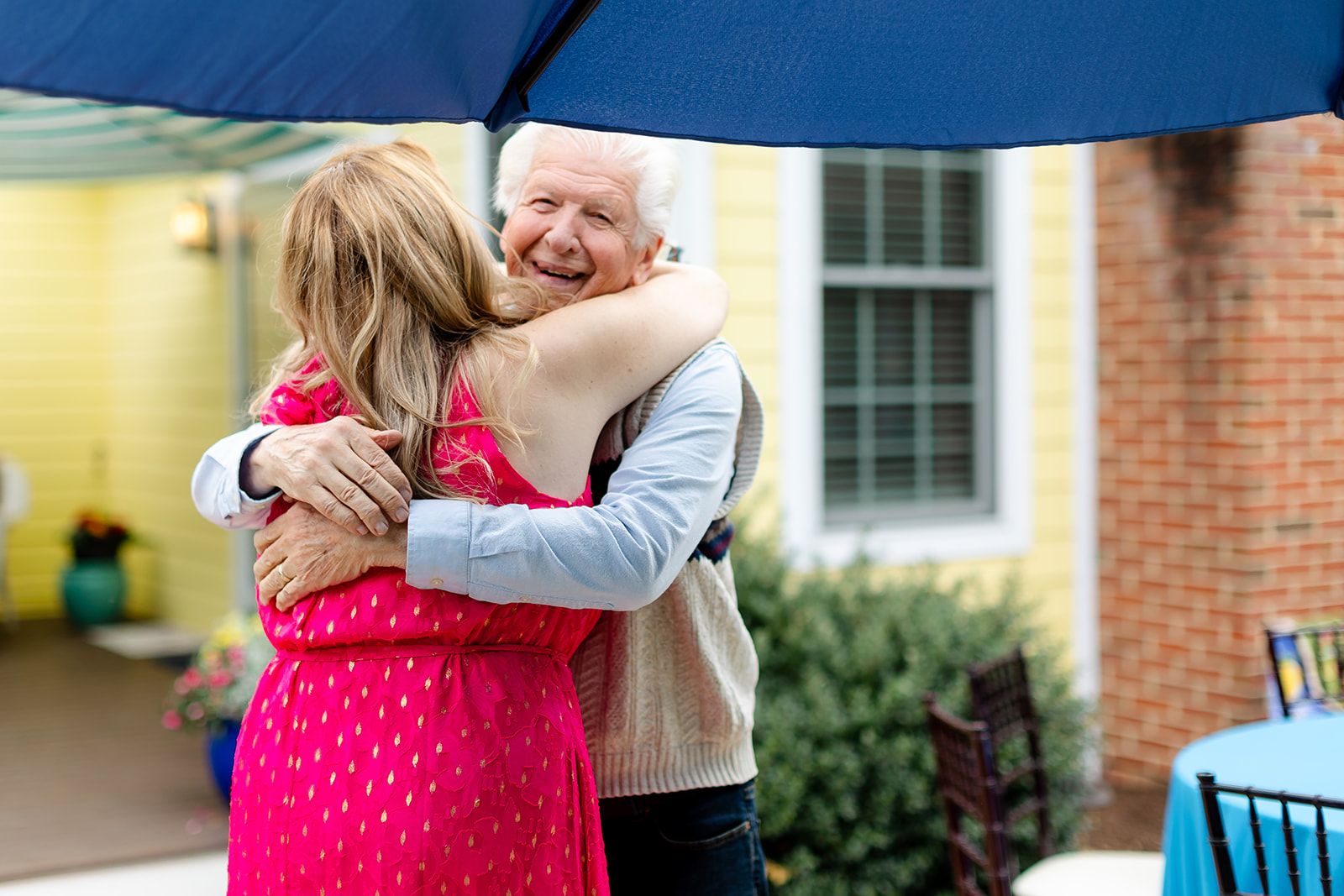 A woman in a bright pink dress hugs a smiling white-haired man in a sweater vest under a large navy umbrella outside a yellow house.