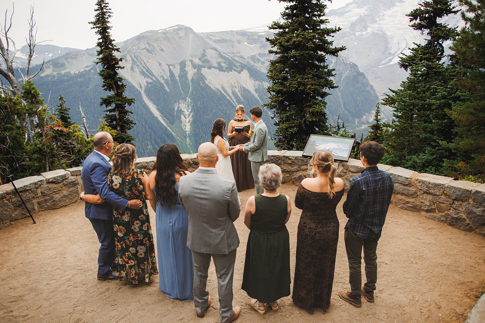 A small group of guests stands in a half circle with their backs to the camera as the couple joins hands in front of an officiant on a stone mountain overlook, one man with his arm around the woman beside him.
