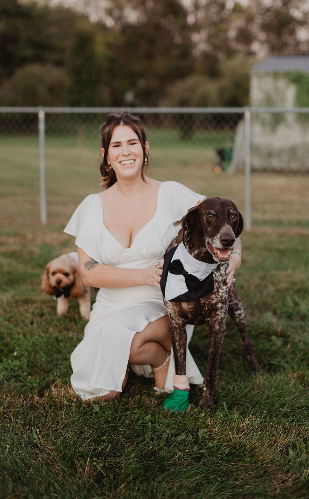 A woman in a white dress crouches in the grass grinning at the camera with her arm around a big brown speckled dog in a tuxedo bib while a small tan dog in a bow tie stands at her side.