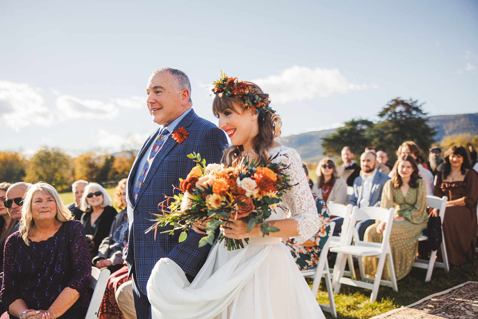 An older man in a navy suit walks the bride down the grassy aisle past seated guests as she smiles wide in a flower crown with a large autumn bouquet.