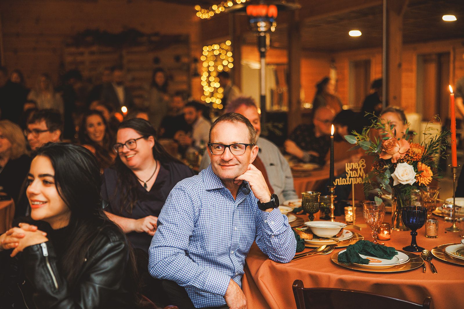 Guests at a reception table smile at something off to the side, one man resting his chin on his hand under warm string lights.