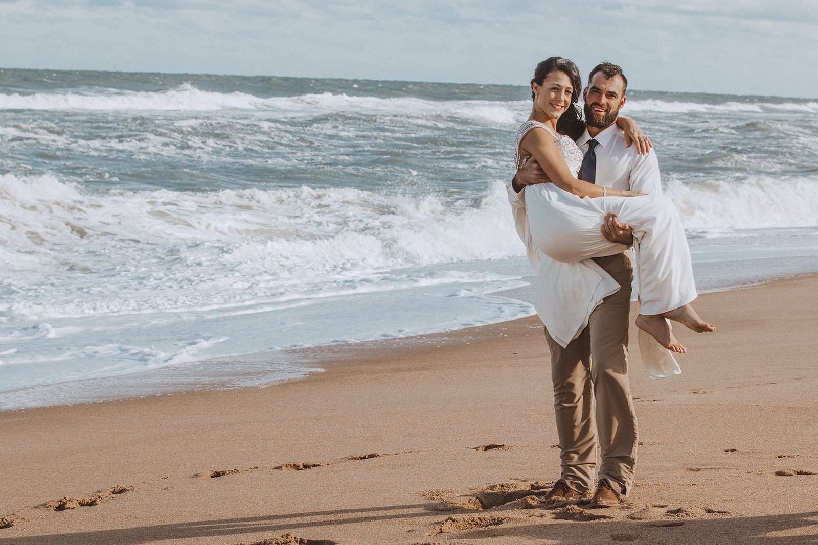 A groom carrying his bride along the surf line on a wide sand beach