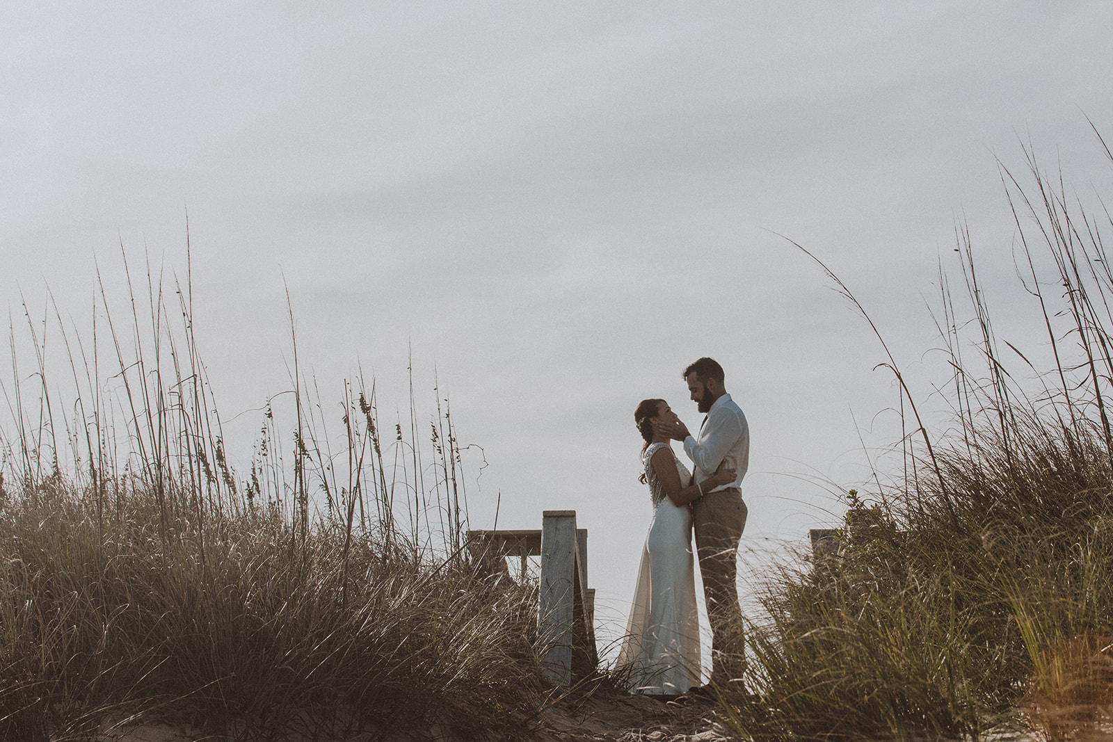 A couple holding each other in the dunes, framed by tall sea oats under a pale sky