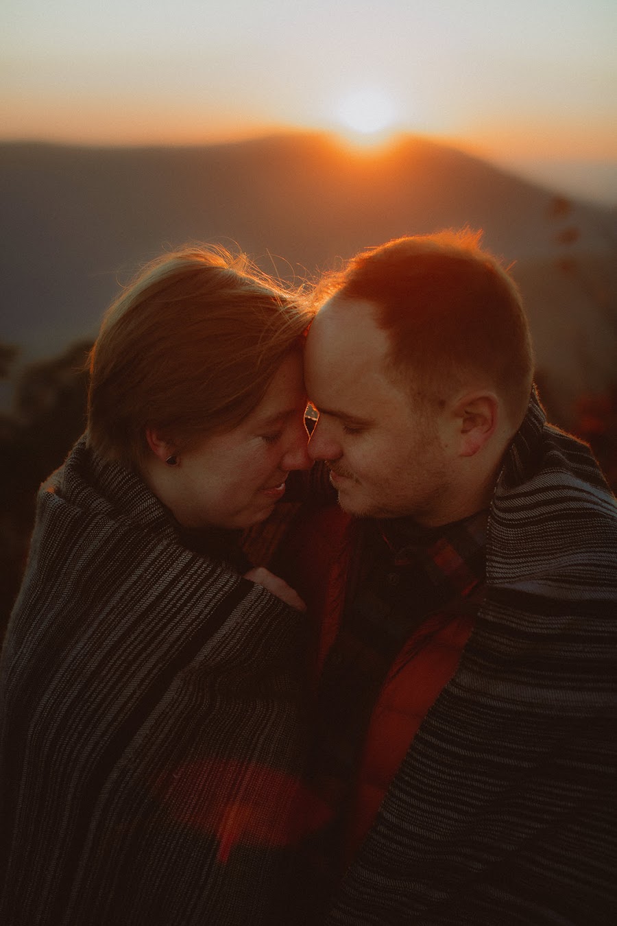 A couple wrapped together in a blanket, forehead to forehead as the sun rises behind the ridge