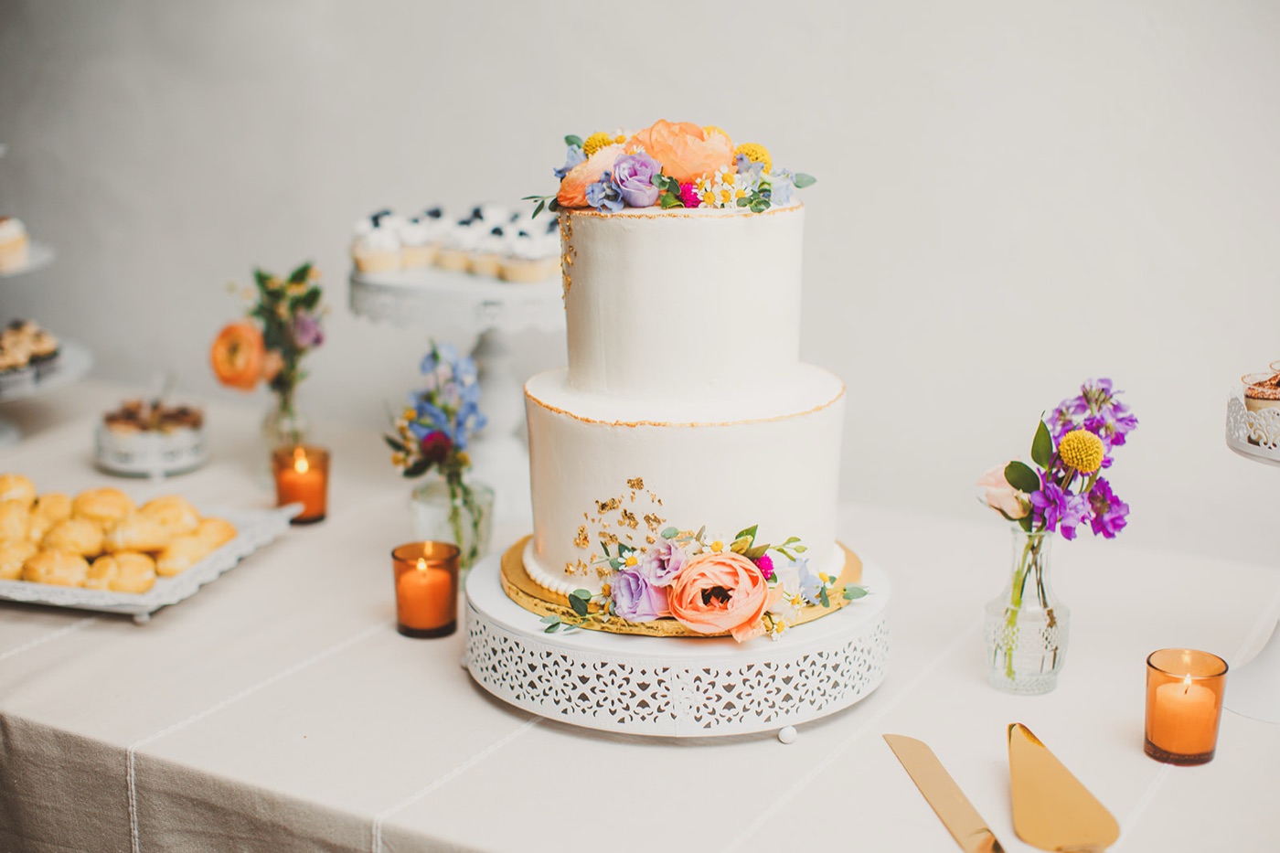 A two-tier white wedding cake with gold-leaf detailing and fresh wildflowers on a filigree stand, amber votives around it